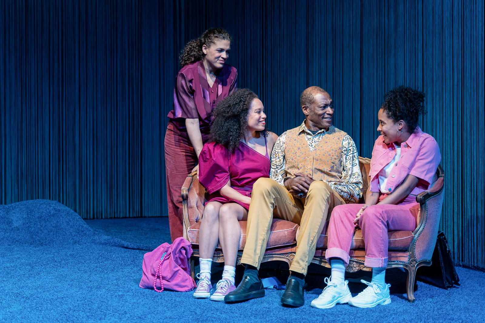 Two women and a man are sitting on a sofa. A woman stands next to it
