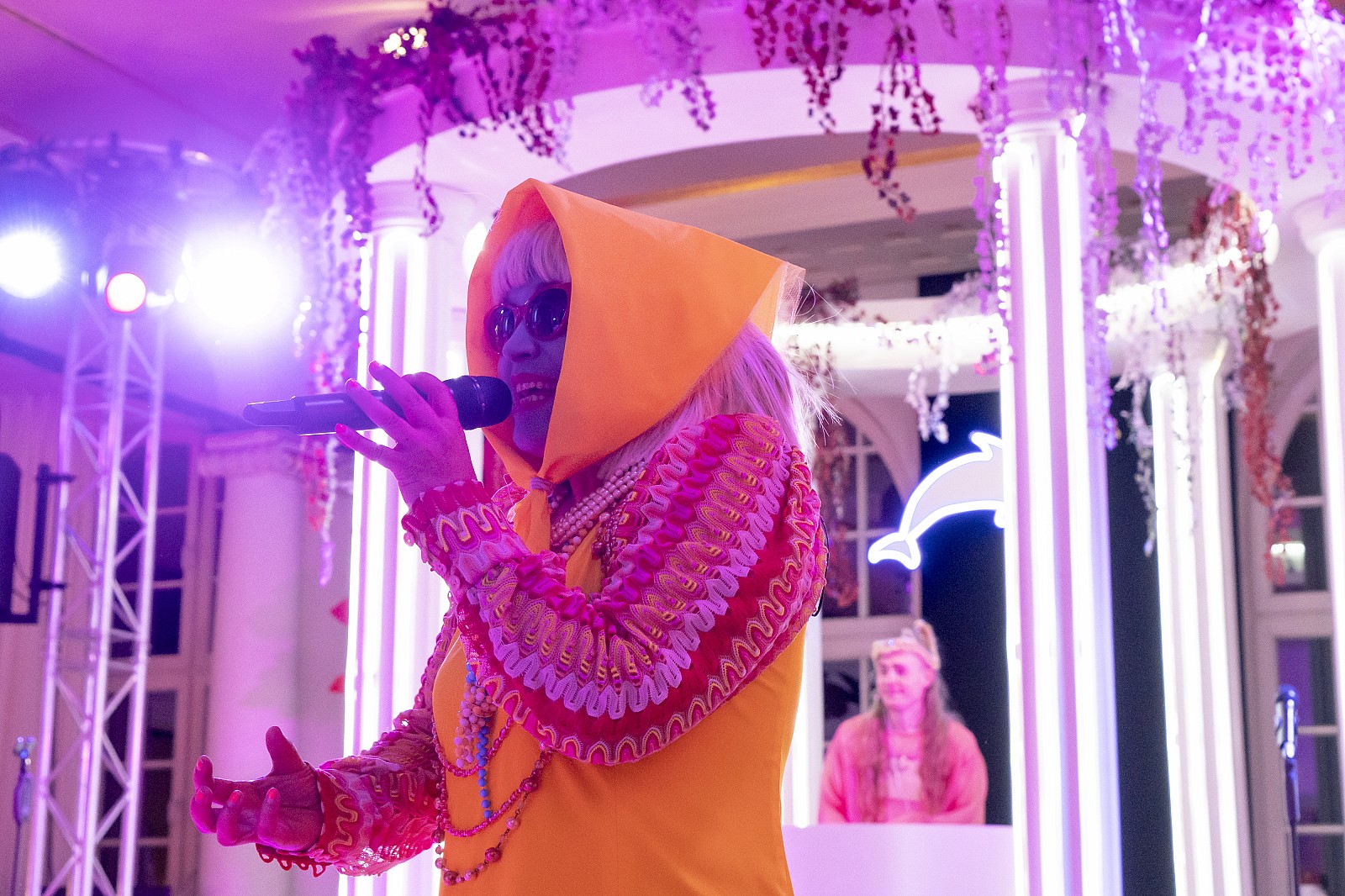 Eine Schauspielerin mit greller Schminke und gelbem Kostüm spricht vor dem DJ Tempel ins Mikrophon.