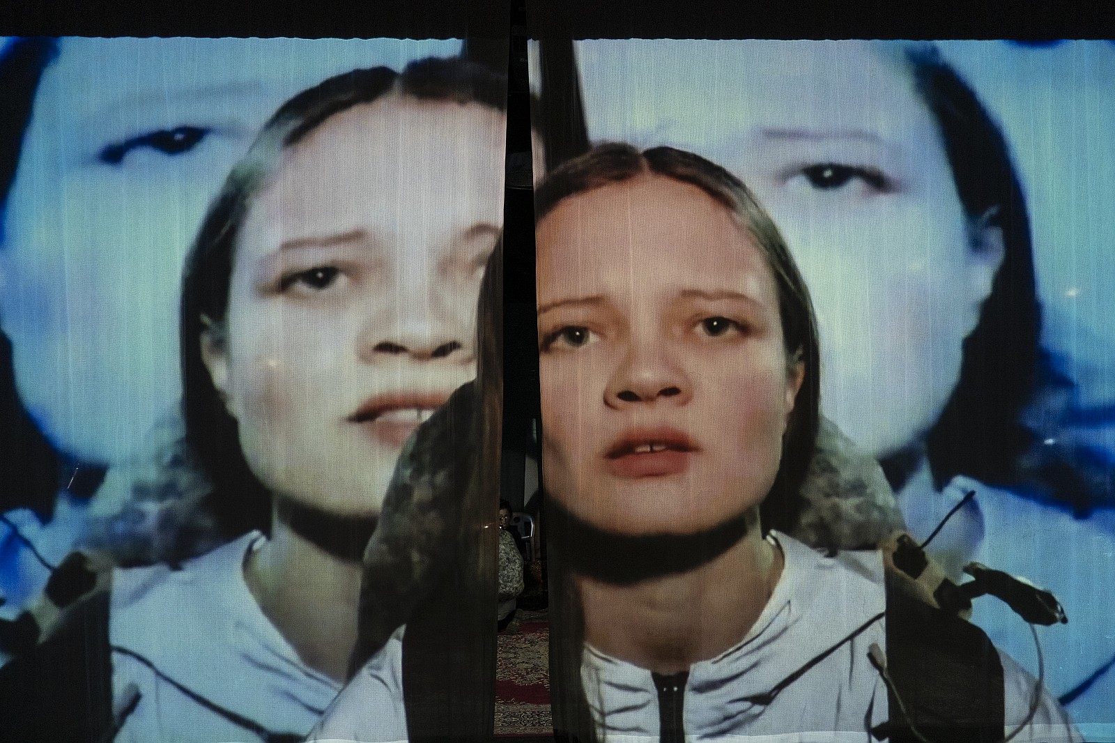 Dreifach hintereinander projiziert erscheint das große Gesicht einer Schauspielerin auf einem Vorhang.